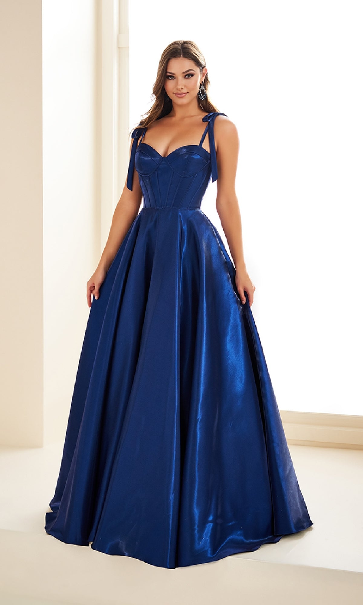 A woman poses indoors in the Ellie Wilde EW36210 Shoulder-Bows Prom Ball Gown, a floor-length deep blue satin A-line dress with a fitted bodice and tied shoulder bows.